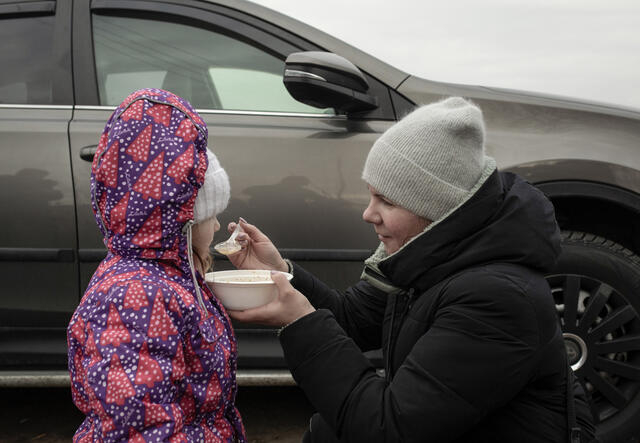 A mother kneels down to feed her daughter, both are outside wearing winter clothes
