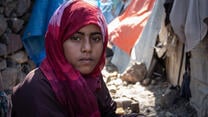 An eleven-year-old girl looks at the camera while sitting outside in a camp for internally displaced people in Yemen. Laundry hangs to dry behind her.